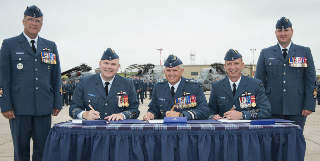 Col Peter Allan, outgoing 12 Wing commanding officer (seated, left); MGen Christian Drouin, Commander of 1 Canadian Air Division; and Col Sid Connor sign the official documents allowing Col Connor to take command of the wing, while CWO Jacques Boucher, 1 Canadian Air Division CWO (standing, left), and 12 Wing CWO David Hepditch (standing, right) observe during the Change of Command Parade at 12 Wing Shearwater, N.S., on July 19, 2017. Cpl Jennifer Chiasson Photo