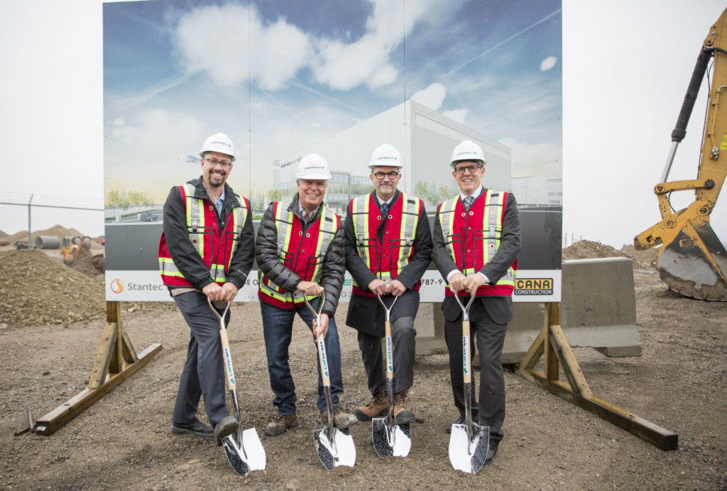 Four men stand with shovels in ground.