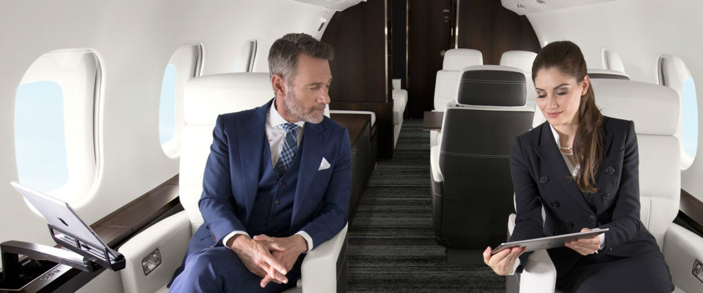 Man and woman sit in the cabin of a business jet, with woman showing man something on a tablet computer.