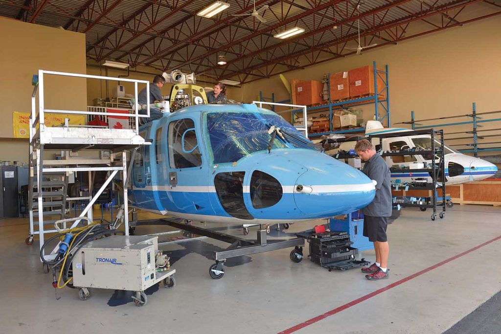 S-76 helicopter rests in a hangar with three men working on it.