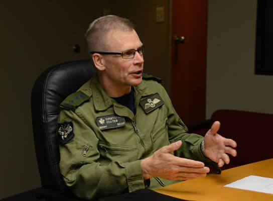 Coates sits at table, speaking, wearing green military uniform
