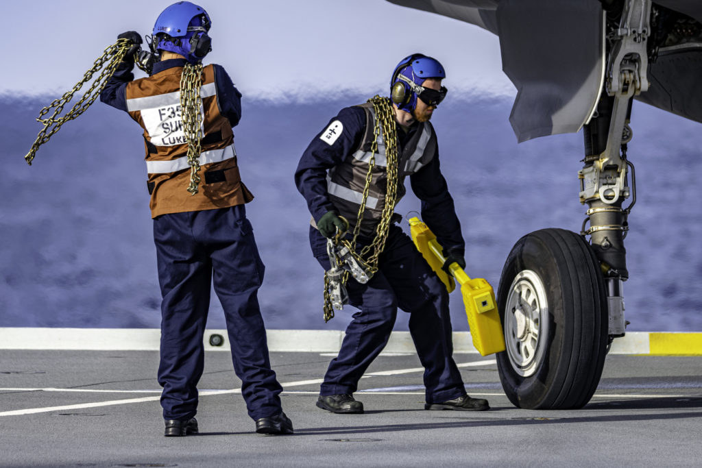 F-35 Mechanics remove chains and wheel chocks in preparation for takeoff. On a flight deck, personell wear different colored jerseys to signify different jobs. These sailors wear brown and gray jersey colors signifing them as Mechanical Engineers. Matt Haskell Photo